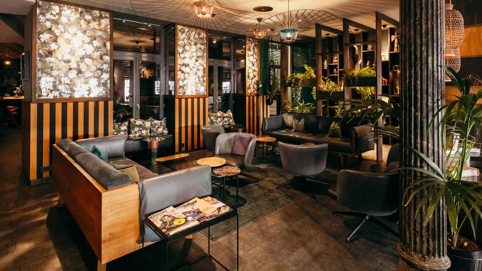 Gemütlicher Lounge- und Barbereich mit dunklen Sofas und Sesseln um niedrige Beistelltische, warm beleuchtet durch mehrere Hängelampen. Im Hintergrund stehen Pflanzenregale und Trennwände mit floral gemusterten Paneelen vor großen Glasfenstern und Türen.