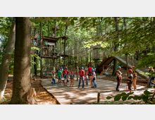 Ein Kletterpark in einem Wald mit Plattformen, Seilbrücken und Kletternetzen zwischen den Bäumen. Auf einer Holzplattform stehen Erwachsene und Kinder mit Kletterausrüstung, begleitet von Betreuern.