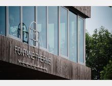 Nahaufnahme eines modernen Gebäudes mit einer Fassade aus Holzpaneelen und großen Glasfenstern. Auf der Holzverkleidung befindet sich das Schild „Fontane Therme Neuruppin“ sowie ein rundes Logo, im Hintergrund sind Bäume sichtbar.