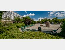 Ein öffentlicher Platz mit gepflastertem Boden, grünen Sitzbänken und mehreren Steinblöcken in der Mitte, umgeben von Bäumen und Sträuchern. Im Hintergrund sind Wohngebäude und ein moderner Gebäudekomplex zu sehen, bei klarem Himmel mit wenigen Wolken.
