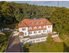 Luftaufnahme eines großen weißen Herrenhauses im klassischen Stil mit roten Ziegeldächern und mehreren Dachgauben, umgeben von einem bewaldeten Gebiet. Vor dem Gebäude befindet sich eine großzügige Terrasse mit Sitzgelegenheiten und Sonnenschirmen sowie eine begrünte Rasenfläche.