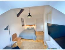 Kleines Schlafzimmer mit weiß gestrichenen Wänden und Holzboden. Ein Bett mit grauer Bettdecke steht an der Rückwand unter einer wandmontierten Lampe, daneben ein Holzstuhl, eine Stehlampe und ein großer Fernseher an einer Seitenwand.