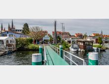 Steg mit grüner Plattform, der zu einem kleinen Hafendorf mit Bootsanlegestellen führt. Im Hintergrund sind Wohnhäuser mit roten Dächern, ein blühender Baum, eine Metallskulptur und Kirchtürme sichtbar.