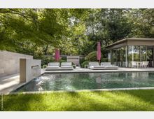 Moderner Außenbereich mit einem rechteckigen Pool, umgeben von einer Terrasse mit weißen Sonnenliegen und rosa Sonnenschirmen. Im Hintergrund ein gläserner Pavillon und dichter Baumbestand, der Schatten spendet.