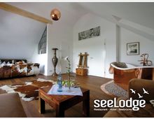 Ein stilvoll eingerichtetes Zimmer mit Holzboden, einer freistehenden Kupferbadewanne, einem rustikalen Holztisch mit Wasserflasche und Dekoration sowie einer Couch mit Kuhfellbezug. Die weißen Wände, schräge Decken und Dachfenster schaffen eine helle, geräumige Atmosphäre.