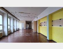 Langer, leerer Flur in einem älteren Gebäude mit großen Fenstern und Heizkörpern auf der linken Seite sowie mehreren Türen entlang des Gangs. Die Decke zeigt abblätternde Farbe und der Boden ist mit Farbresten und Schmutzspuren bedeckt; rechts sind gelb gestrichene Wände mit verblasster Wanddekoration zu sehen.