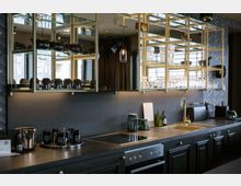 Moderne Küchenzeile mit dunklen Schränken, goldenen Griffen und einer Arbeitsplatte aus Stein. Darüber befinden sich offene, goldgerahmte Regale mit Gläsern, Geschirr und Dekorationselementen, sowie ein integrierter Spiegel und Hängelampen.