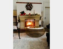 Ein gemütliches Wohnzimmer mit einem Kamin aus beigem Stein, der von Weihnachtsdekorationen wie einem Kranz und Girlanden mit Lichtern geschmückt ist. Im Vordergrund steht ein runder, goldener Metalltisch auf einem hellen Teppich.