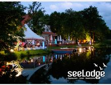 Nachtaufnahme eines eleganten Restaurants am Ufer eines Sees, umgeben von Bäumen. Der Außenbereich ist mit einer Terrasse, weißen Sonnenschirmen, bunten Lichtern und Booten dekoriert, die sich im ruhigen Wasser spiegeln.