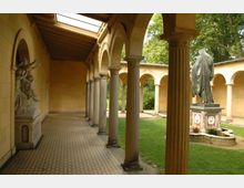 Überdachter Arkadengang mit Säulen aus Stein und einem Mosaikboden, daneben ein Innenhof mit Rasen und einer Statue auf einem verzierten Sockel. Im Hintergrund sind grüne Bäume und weitere Arkaden zu sehen.