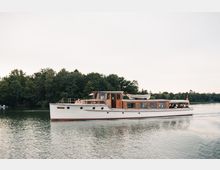 Ein weißes Ausflugsschiff mit Holzelementen fährt auf einem ruhigen See, umgeben von dicht bewaldeten Ufern. Das Boot trägt ein auf dem Dach befestigtes Beiboot und ist mit mehreren Fenstern ausgestattet, hinter denen Passagiere sichtbar sind.