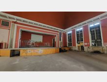 Ehemaliger Veranstaltungssaal mit Bühne, abblätternder roter Wandfarbe und großen Fenstern mit beschädigten Rahmen. Der Raum zeigt Graffiti an den Wänden und weist sichtbare Spuren des Verfalls auf.