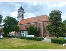 Backsteinkirche mit Satteldach und einem hohen Turm mit geschwungener Haube, umgeben von Bäumen und einer Grünfläche; im Vordergrund ein Weg sowie geparkte Fahrzeuge.