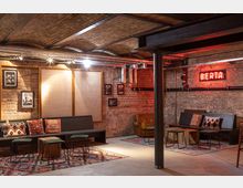 Industriell gestalteter Raum mit freiliegenden Ziegelwänden, sichtbaren Rohren an der Decke und gedämpfter Beleuchtung. Sitzbereich mit Holzbänken, bunten Kissen sowie Teppichen im Boho-Stil, dazu eine rote Neonleuchtschrift mit der Aufschrift „Berta“.