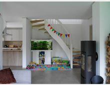 Wohnraum mit einer weißen Wendeltreppe in der Mitte, darunter ein Aquarium und ein Kinderbereich mit bunten Spielsachen und einer Spielmatte. Rechts steht ein schwarzer Holzofen, daneben gestapeltes Brennholz, und links ist eine offene Küche mit weißen Schränken und Holzarbeitsfläche sichtbar.