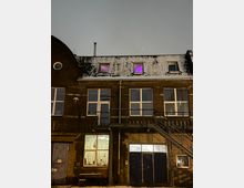Backsteinfassade eines mehrstöckigen Gebäudes mit großen Fenstern, einer Außentreppe auf der rechten Seite und Schnee auf dem Dach. Zwei Fenster im Dachgeschoss leuchten in violettem Licht, während im Erdgeschoss ein beleuchteter Raum mit Vorhängen zu sehen ist.