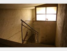 Ein einfaches, heruntergekommenes Treppenhaus mit bröckelnden Wänden, einem Metallgeländer und einem großen Fensterausschnitt, durch den diffuses Tageslicht einfällt.