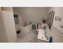 Badezimmer mit einer Eckbadewanne, einem Doppelwaschbecken mit großem Wandspiegel, einer Toilette und mehreren weißen Schränken. Der Raum hat eine schräge Decke, eine Fensterjalousie, Dekorationen und Handtücher in Blau- und Türkistönen.