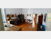 Wohnküche mit Holzboden und weißen Wänden, ausgestattet mit einem schwarzen Kühlschrank, einem Gasherd, offenen Holzregalen und einer Spüle auf einer Arbeitsplatte. Im Hintergrund ein kleiner Esstisch mit zwei Stühlen, sowie eine Tür zum angrenzenden Raum.