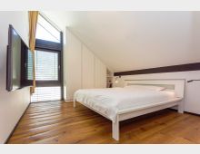 Modernes Schlafzimmer mit großem weißen Bett auf Holzdielenboden, Einbauschrank unter einer schrägen Decke und Wandregal mit Leselampe. Links ein großes Fenster mit Jalousien und Vorhängen sowie ein an der Wand montierter Fernseher.