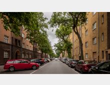 Wohnstraße mit mehrstöckigen, gelb-braunen Gebäuden auf beiden Seiten, flankiert von Bäumen, deren Äste die Straße überspannen. Autos sind parallel entlang der Bordsteine geparkt, und die Straße führt zentral in die Ferne.