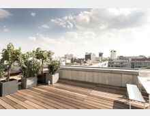 Dachterrasse mit Holzbodenbelag, bepflanzt mit mehreren Kübelpflanzen, darunter ein kleiner Baum, und einer Sitzbank. Im Hintergrund ist eine urbane Skyline mit modernen Gebäuden und klarem Himmel zu sehen.