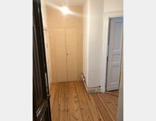 Ein schmaler Flur mit hellem Holzdielenboden und weißen Wänden. Rechts führt eine Tür mit klassischen Zierleisten in einen angrenzenden Raum, links befindet sich ein dunkler Türrahmen, und am Ende des Flurs ist eine Einbauschranktür aus hellem Holz zu sehen.