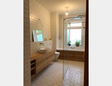 Helles Badezimmer mit weißen Fliesen an den Wänden und dem Boden. Ein Holzwaschtisch mit Aufsatzwaschbecken steht links, eine Glasdusche und eine Badewanne aus Holzoptik befinden sich im Hintergrund; vor dem Fenster stehen Pflanzen.