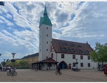 Weitläufiger Platz mit einer historischen Kirche, die einen hohen weißen Turm mit grünem Spitzdach und einer Uhr besitzt. Das Kirchenschiff hat ein rotes Ziegeldach mit Dachfenstern; im Vordergrund befinden sich Bäume, Lampen und mehrere Fahrräder.