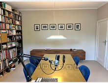 Ein Arbeitszimmer mit einem großen goldenen Tisch, blauen Stühlen und einem Mikrofon-Setup. An der Rückwand stehen ein Holz-Sideboard, fünf gerahmte Schwarz-Weiß-Fotografien und ein großes Bücherregal gefüllt mit Büchern.