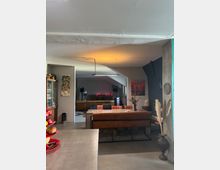 Ein modernes Wohnzimmer mit offener Gestaltung, das von einer Mischung aus industriellen und gemütlichen Elementen geprägt ist. Im Vordergrund befinden sich eine Küchenarbeitsplatte und eine Obstschale, während im Hintergrund eine braune Ledercouch, ein Tisch mit einem Laptop und dekorative Elemente wie Bilder und Trockenblumen zu sehen sind.