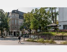 Straßenszene mit einem Eckgebäude im Altbaustil, das Geschäfte im Erdgeschoss enthält, darunter ein Friseursalon und eine Bäckerei. Rechts daneben befindet sich eine Terrasse mit Tischen und Sonnenschirmen, umgeben von Bäumen und Grünflächen; im Vordergrund ist ein Fußgänger zu sehen.