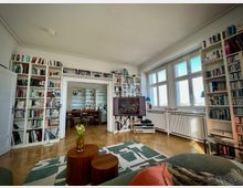 Heller Wohnraum mit hohen Decken, großen Sprossenfenstern und Parkettboden, umgeben von deckenhohen weißen Bücherregalen. In der Mitte stehen ein Teppich und zwei runde Holztische; durch eine breite Durchgangsöffnung ist ein angrenzendes Esszimmer sichtbar.