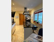 Moderne Küche mit cremefarbenen Wänden und gefliesten Böden. Ein zentraler Küchenblock mit schwarzer Arbeitsfläche und einer hängenden Edelstahl-Dunstabzugshaube; auf der rechten Seite ein Fenster mit Blumenkästen. Im Hintergrund eine Holztür und Kühlschrank mit Magneten.