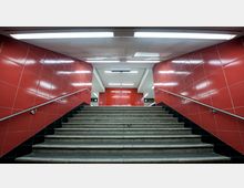 Treppenaufgang in einer U-Bahn-Station mit roten gefliesten Wänden und silbernen Handläufen auf beiden Seiten. Die Decke ist weiß mit integrierter Beleuchtung, und oben sind Beschilderungen mit Informationen sichtbar.