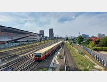 Stadtansicht mit einer vorbeifahrenden S-Bahn auf mehrgleisigen Bahngleisen, flankiert von einer großen Einkaufsmall auf der linken Seite und Graffiti-verzierten Zäunen auf der rechten Seite. Im Hintergrund sind Hochhäuser, Kräne und der Berliner Fernsehturm sichtbar.