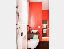 Modernes Badezimmer mit einer auffälligen roten Wand, einer Wandtoilette, einem Waschbecken auf einem dunklen Waschtisch sowie weißen Fliesen an der Duschwand. Im Hintergrund befinden sich schwarze Handtuchhalter mit grauen Handtüchern.