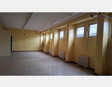 Leerer, länglicher Innenraum mit gelb gestrichenen Wänden, mehreren hohen Fenstern in tiefen Nischen und Heizkörpern darunter. An der Decke verlaufen Rohrleitungen und lange Leuchtstofflampen über einem grauen Boden.