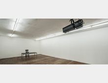 Ein leerer, rechteckiger Raum mit weißen Wänden und Holzfußboden. In der Mitte stehen ein schlichter Tisch und ein Stuhl, an der Decke befinden sich lange Neonleuchten sowie ein montierter Scheinwerfer.