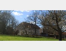 Altes Landhaus mit rotem Ziegeldach, umgeben von kahlen Bäumen auf einer Wiese, bei sonnigem Wetter unter blauem Himmel. Ein Nebengebäude mit Dachziegeln ist rechts im Hintergrund zu sehen.