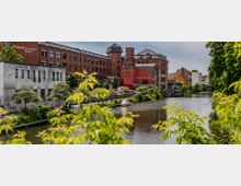 Eine Uferpromenade mit einem ruhigen Fluss, umgeben von Gebäuden, darunter ein rotes Backsteingebäude mit der Aufschrift "Sankt Annen Höfe". Im Vordergrund sind grüne Bäume und Büsche zu sehen, während am Ufer Boote und Spazierwege erkennbar sind.