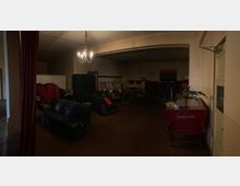 Ein schwach beleuchteter Innenraum mit dunklem Teppichboden, mehreren schwarzen Sofas, roten Vorhängen und einem Kronleuchter an der Decke. Im Hintergrund stehen leere Stühle, Bilderrahmen und Stapel von Gegenständen; rechts ist ein roter Eiswagen mit der Aufschrift "Carte d'Or" zu sehen.