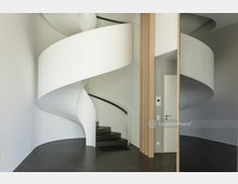 Moderner Innenraum mit einer weißen, geschwungenen Wendeltreppe, dunklem Bodenbelag und glatten, hellgrauen Wänden; eine schlichte Tür und vertikale Holzpaneele befinden sich rechts.