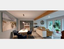 Helles Wohnzimmer und Essbereich mit moderner Einrichtung. Links steht ein Esstisch mit blauen Stühlen vor einer Wand mit Strandbild, rechts befinden sich eine helle Couch, eine Zimmerpflanze und große Fenster, die Blick ins Grüne bieten.