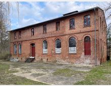 Zweistöckiges Backsteingebäude mit großen, bogenförmigen Fenstern im Erdgeschoss und kleineren rechteckigen Fenstern im Obergeschoss. Das Gebäude hat rote Türen, bröckelnde Fassade und steht in einer bewachsenen Umgebung mit freier, befestigter Fläche davor.