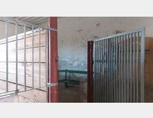 Innenansicht eines einfachen Stalls mit Metallgittern, Holzwänden und einem abgenutzten Putz an der Ziegelwand im Hintergrund. Am Boden sind Heu und Staub sichtbar, links steht eine hölzerne Abtrennung, und im Hintergrund befindet sich ein grüner Gestellrahmen.