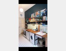 Küche mit einer Arbeitsplatte aus Holz, unter der ein Herd, eine Spülmaschine und eine Waschmaschine integriert sind. An der dunkelblau gestrichenen Wand hängen Regale mit Küchenutensilien, darüber befinden sich drei bunte Gemälde; die Rückwand ist mit hellblauen Fliesen verkleidet.