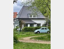 Einfamilienhaus mit weißer Fassade und dunklem Schrägdach, umgeben von Bäumen und Sträuchern; im Vordergrund ein hellblauer Volkswagen Käfer auf einer Grünfläche mit Wegen.