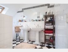 Badezimmer mit zwei weißen Standwaschbecken, großem Wandspiegel und schwarz-weiß gefliestem Boden. Rechts steht ein Holzregal mit Handtüchern, links sind ein kleines Fenster und ein Handtuchhalter sichtbar.