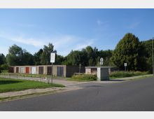 Reihen von einfachen Garagen mit unterschiedlich farbigen Türen stehen an einer Straße, umgeben von Grünflächen und Bäumen. Im Vordergrund sind ein Asphaltweg, eine Wiese sowie ein Verkehrsschild und eine Schranke zu sehen.